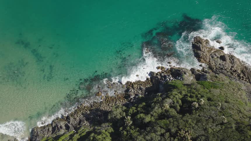 Static aerial top-down shot of beautiful turquoise ocean water crashing against a rocky headland covered in green trees