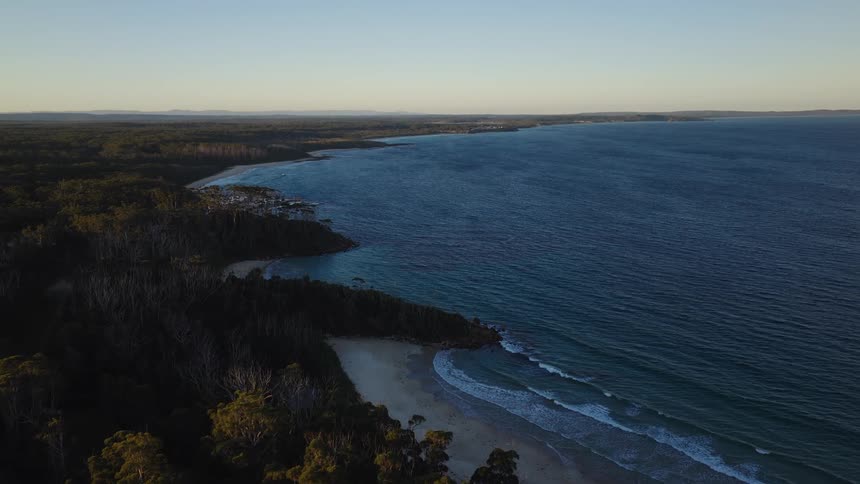 A serene static aerial view captures the vast blue ocean meeting a forested coastline with a sandy beach during a beautiful sunrise