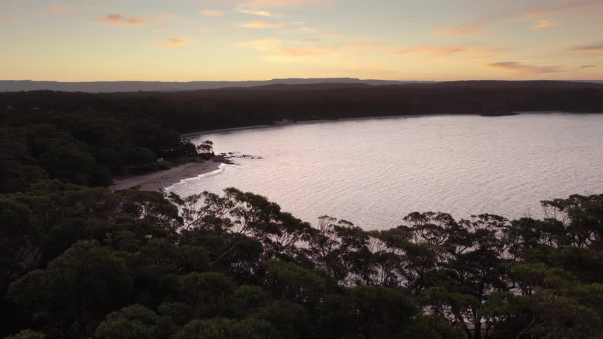 A slow aerial view pushing over a lush coastal forest reveals a secluded bay with a sandy beach during a beautiful sunset