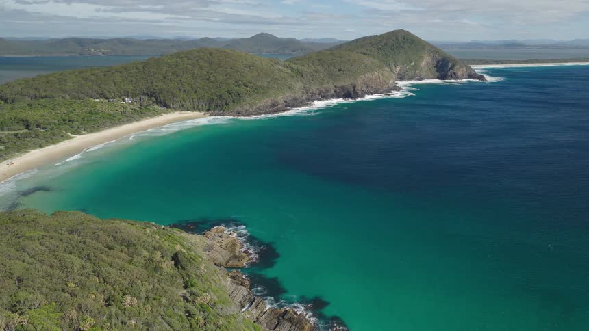 A breathtaking high-angle view captures a secluded sandy beach cove with vibrant turquoise waters along a lush, green coastline