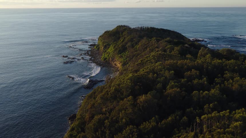 An aerial shot slowly pulls back to reveal a lush, tree-covered headland jutting into the calm ocean waters at sunrise