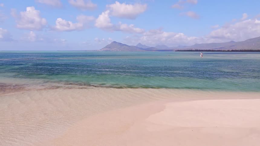 Idyllic tropical beach scene with a pristine sandbank, clear turquoise water, and distant mountains under a blue sky