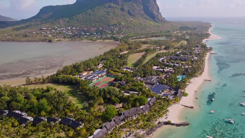An aerial ascent reveals a stunning tropical paradise with a luxury beach resort, turquoise ocean, and a dramatic mountain backdrop in Mauritius