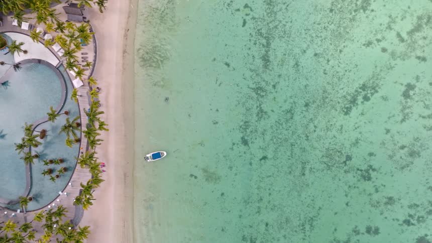 A serene top-down aerial view slowly ascends to reveal a luxurious tropical resort with a pristine beach, palm trees, and a stunning pool next to the clear turquoise ocean