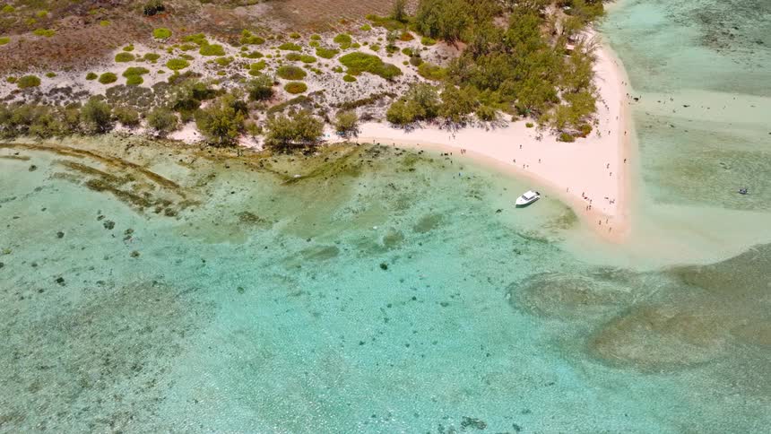 An aerial drone shot slowly pulls back and tilts up to reveal a stunning tropical island with a white sand beach, turquoise water, and a moored boat