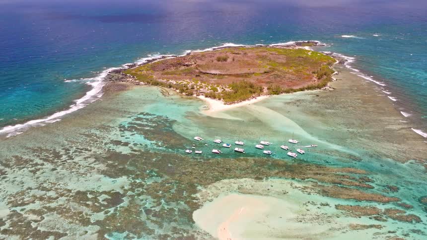 A breathtaking aerial shot tilts up to reveal a stunning tropical island connected by a sandbar, surrounded by turquoise water and anchored boats