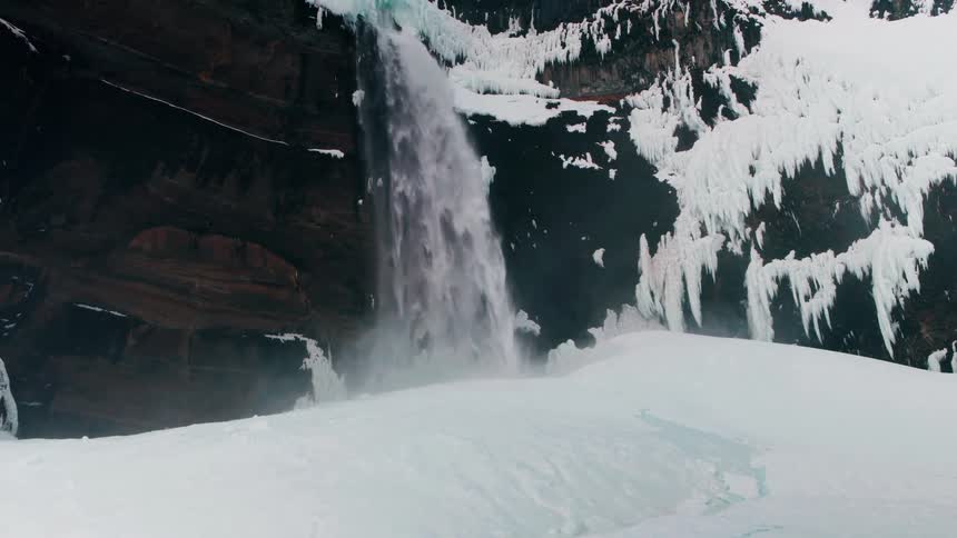 A powerful waterfall crashes down a rocky cliff into a snowy and icy basin during winter