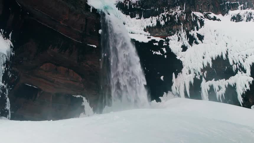 A powerful waterfall crashes into a snowy basin, surrounded by an icy cliff face in a stark winter landscape