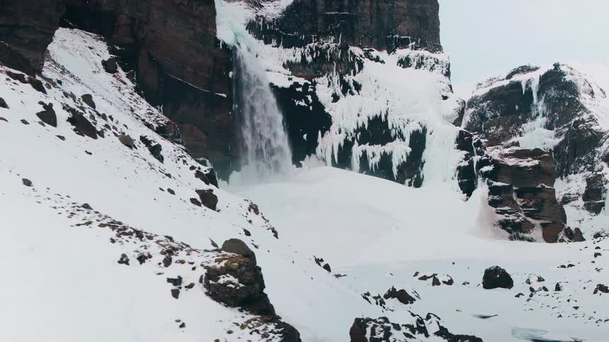 An epic aerial shot pushes in on a massive waterfall cascading through a frozen, snow-covered canyon in Iceland