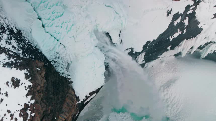 A breathtaking top-down aerial view follows the powerful flow of a partially frozen waterfall as it plunges into an icy basin below