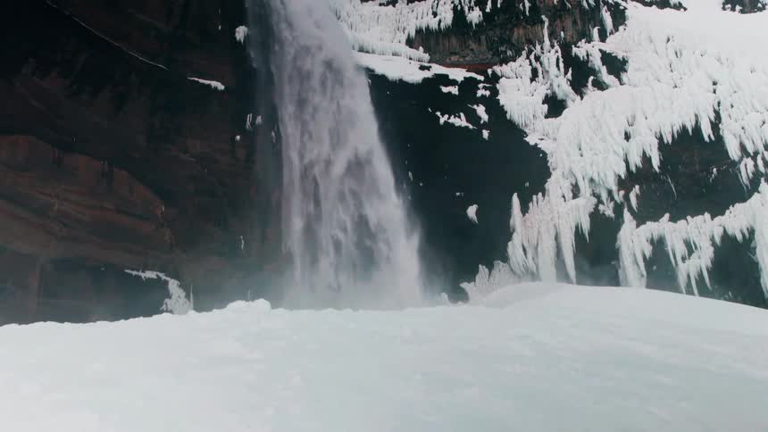 Slow tilt up reveals a powerful waterfall crashing onto a mound of snow with icy cliffs in the background