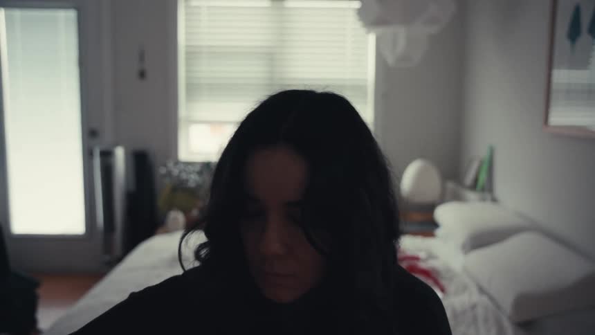 A somber woman with dark hair looks down with her face in shadow while sitting in her bedroom