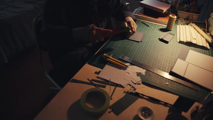 A focused artisan works diligently at a cluttered desk in a dimly lit room, using a small hammer to craft a handmade item