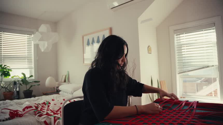 A focused artist weaves red ribbons into an intricate pattern in her sunlit bedroom studio