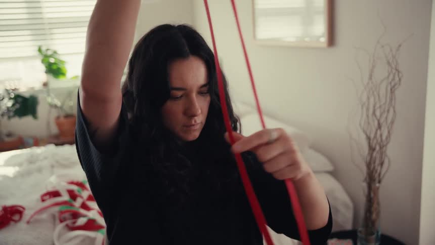 A woman with dark wavy hair carefully measures a long red ribbon while preparing to wrap gifts in her bedroom