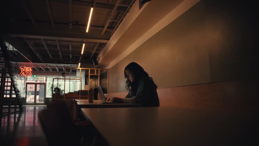 A focused young woman works late on her laptop in a dimly lit, modern, and stylish co-working space