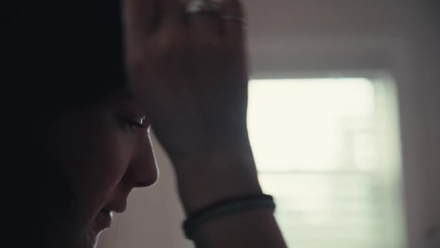 A close-up, silhouetted profile of a young woman brushing her hair away from her face in front of a bright window