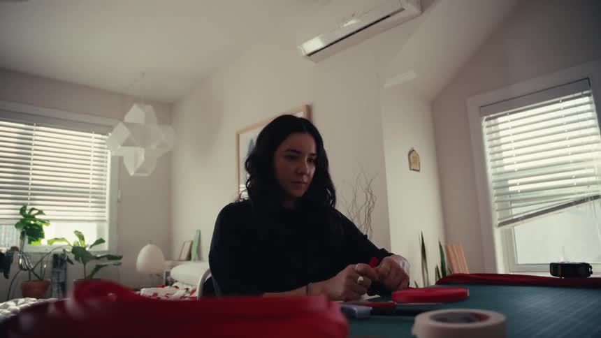 A focused young woman crafts with red material at a table in her sunlit home studio