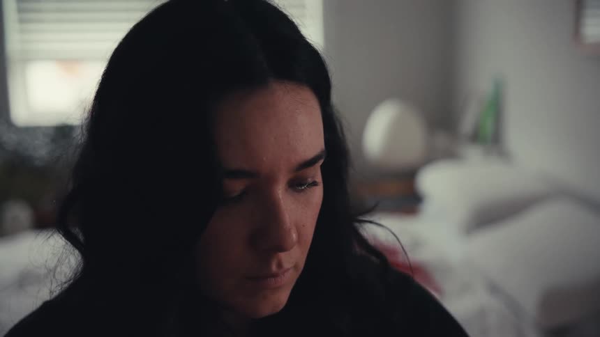 A close-up of a sad and pensive woman with dark hair looking down while sitting in her dimly lit bedroom