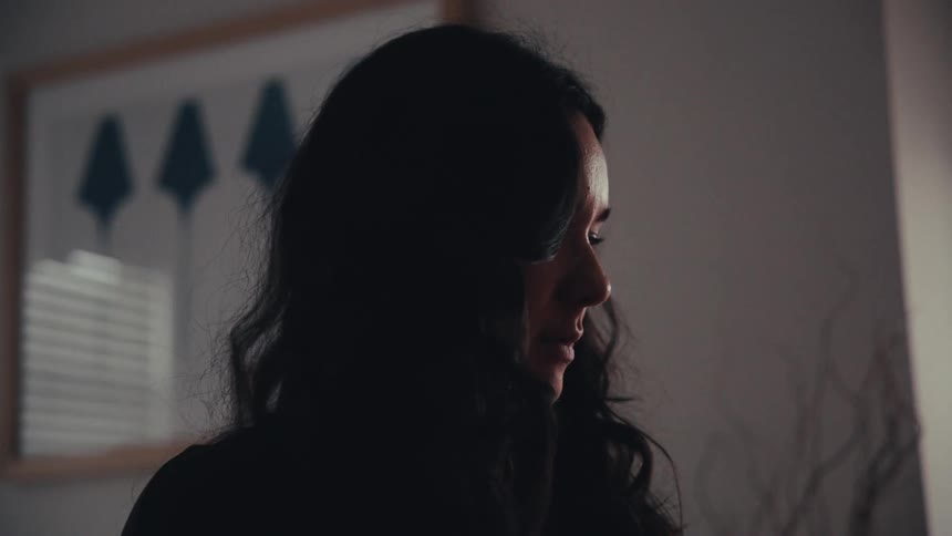 A moody close-up shot captures a woman with long dark hair looking down thoughtfully in a dimly lit room