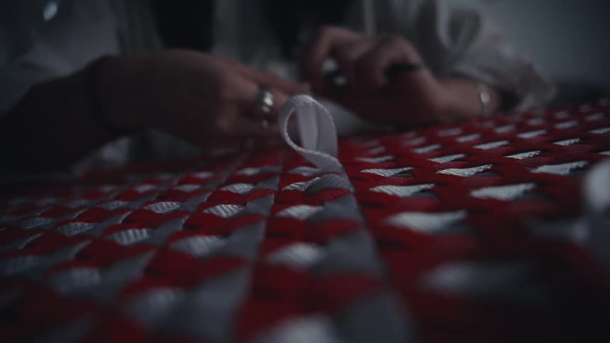 Close up slow motion of hands carefully weaving a white strap through a red and white patterned textile