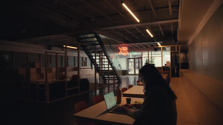 A person works alone on their laptop in a large, dimly lit, modern co-working space