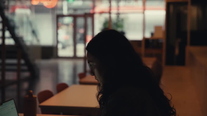 A silhouetted woman works on her laptop in a modern office with a view of the busy city street outside