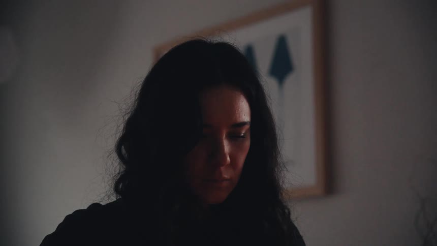 A contemplative woman with dark hair sits alone in a dimly lit room, her face illuminated by a subtle, flickering light