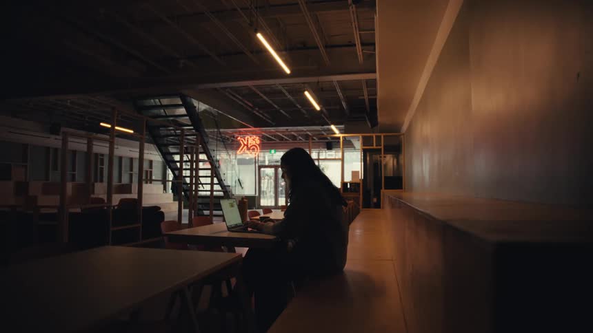 A person works alone on a laptop in a dimly lit, modern co-working space
