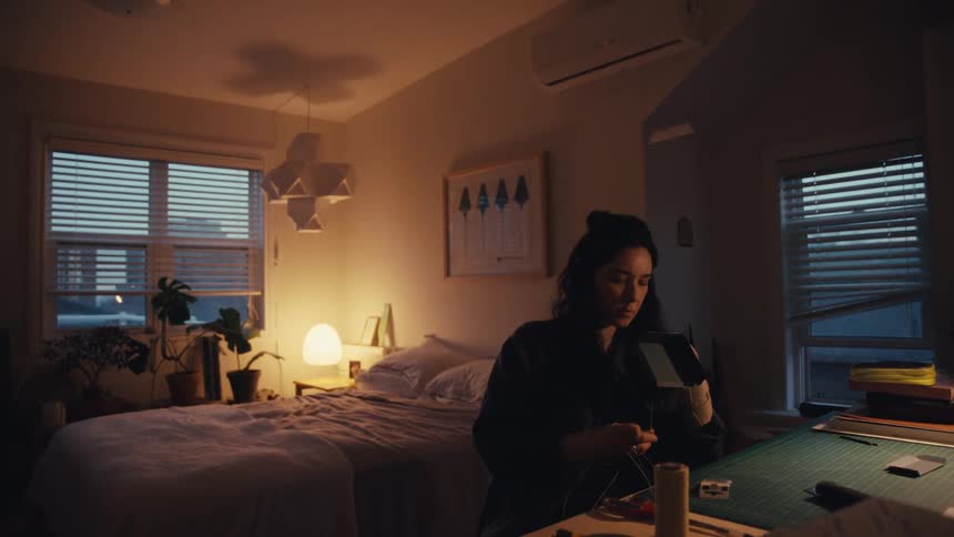 A focused female artist meticulously hand-sews a creative project at her desk in a dimly lit, cozy bedroom