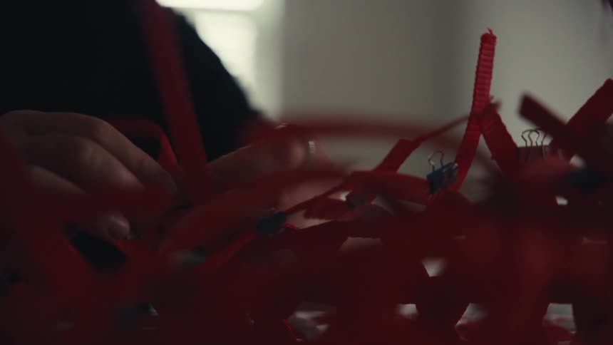 An artist's hands meticulously weave vibrant red ribbons together, creating an intricate pattern in a dimly lit creative space