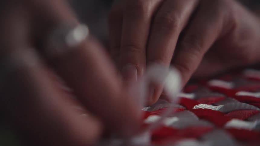 An artisan's hands meticulously weave a white ribbon through an intricate red and grey patterned textile in a slow-motion close-up