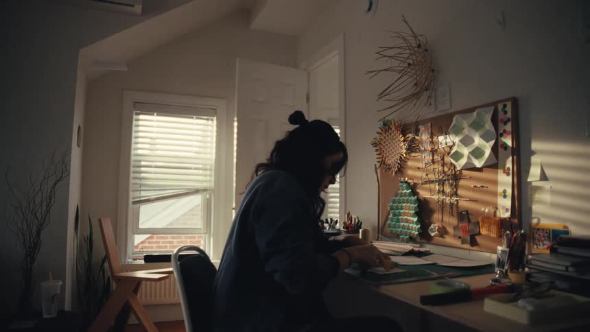 A focused artist sits at their desk in a cozy home studio, working on a project as dramatic sunlight streams through the blinds