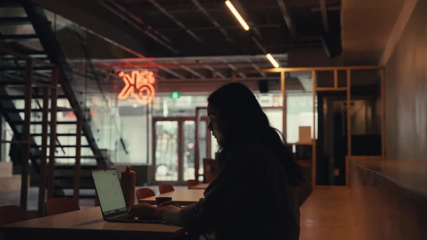 A silhouetted woman with long hair types on her laptop while working late in a moody, industrial-style office