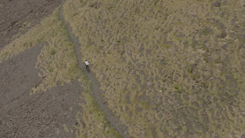 An aerial view captures a lone mountain biker riding along a narrow trail on a vast, rugged mountainside