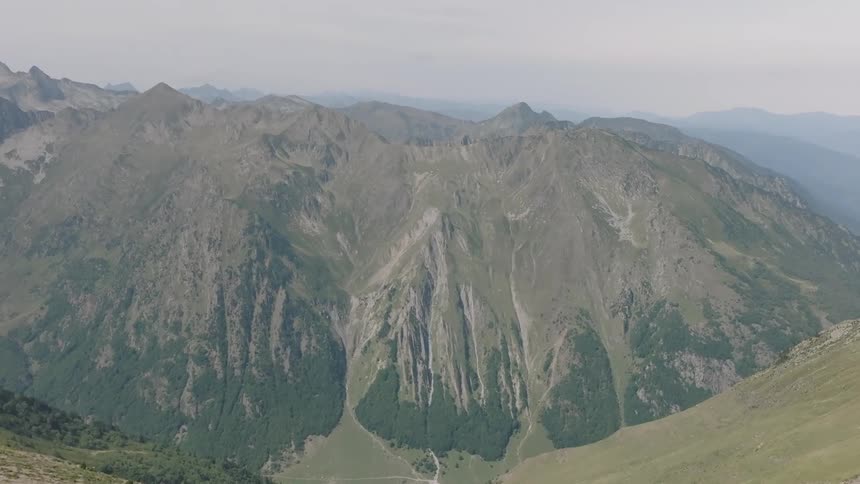 A cinematic aerial shot slowly pushes over a grassy mountain ridge to reveal a breathtaking view of a majestic mountain range
