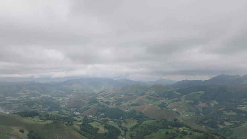 A serene, moody landscape captures rolling green hills and a vast mountain valley under a blanket of gray, overcast clouds