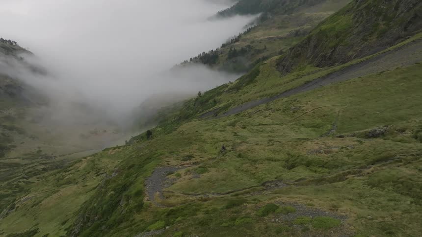 Epic aerial footage shows a mountain biker descending a steep, grassy slope into a valley filled with low-hanging clouds