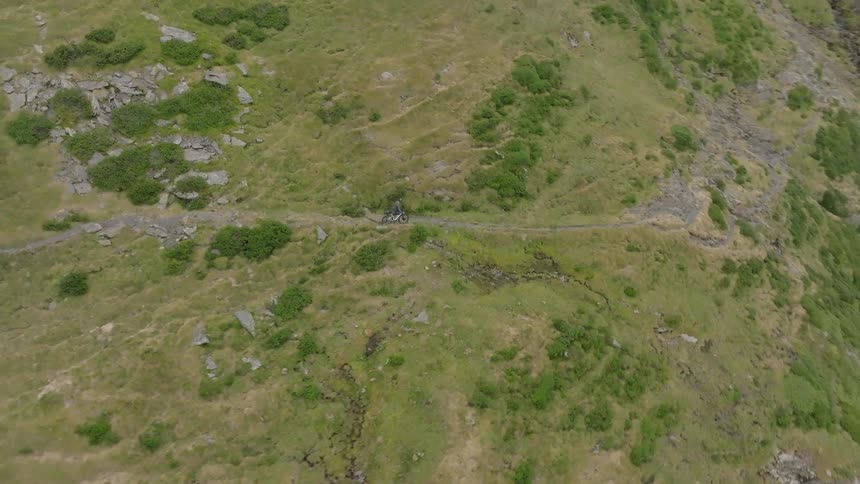 An aerial view captures a mountain biker riding along a narrow, winding dirt trail on a vast, green mountainside