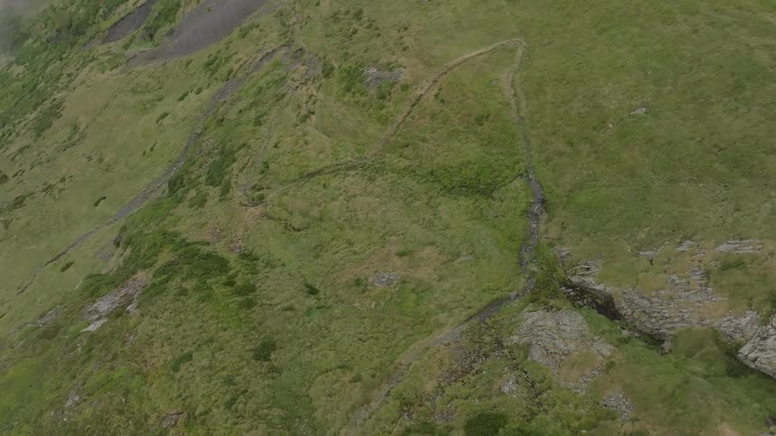 An aerial view follows a lone hiker walking up a winding trail on a vast, green mountainside on a foggy day