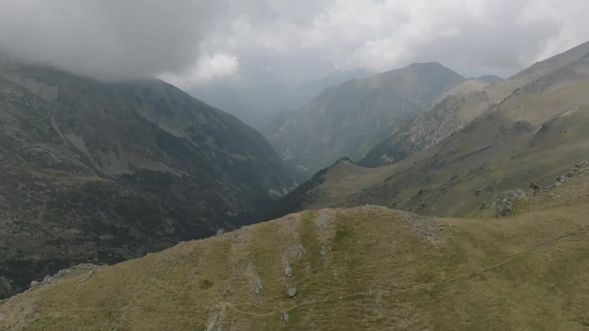 A slow aerial push over a grassy mountain peak reveals a vast, cloudy valley with a small lake below