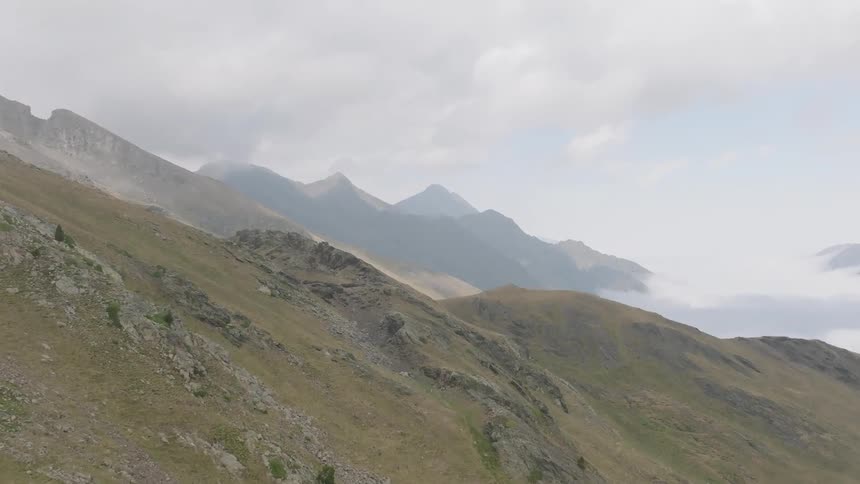 An aerial shot pans across a vast mountain range on an overcast day, revealing a valley filled with a sea of clouds