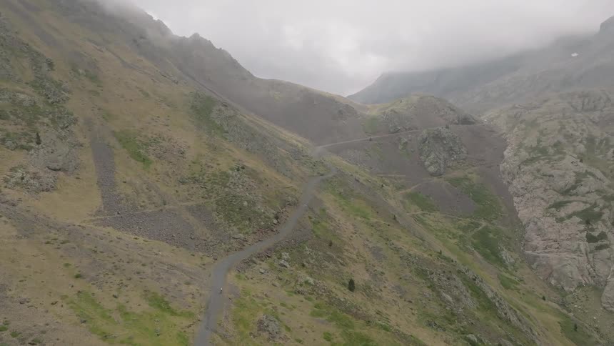 An epic aerial shot follows a lone cyclist navigating a winding dirt road up a massive, cloud-covered mountain