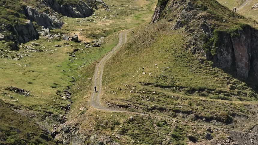 A lone hiker with a backpack walks up a winding dirt trail in a vast, green mountain valley