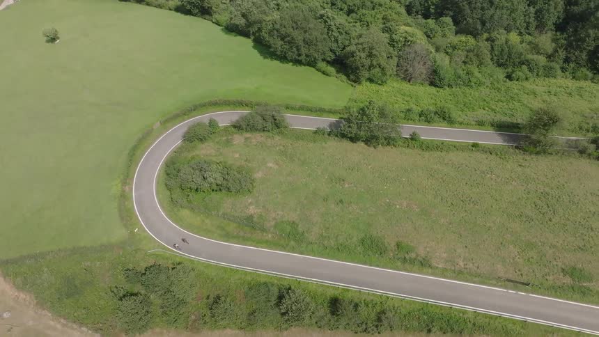 An aerial view shows two cyclists navigating a sharp hairpin turn on a scenic country road surrounded by lush green fields and forests