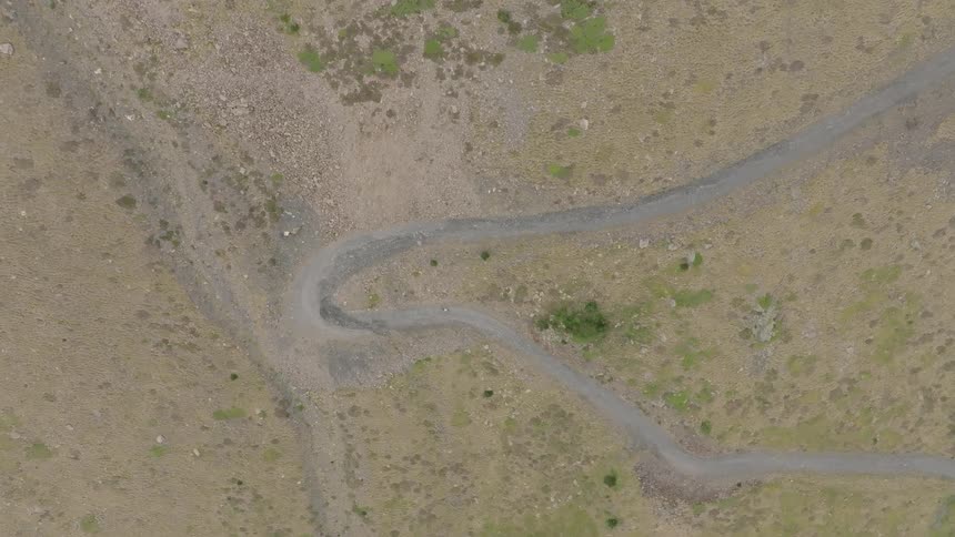 Overhead aerial view of a lone hiker making their way up a winding dirt road on a desolate, grassy mountainside