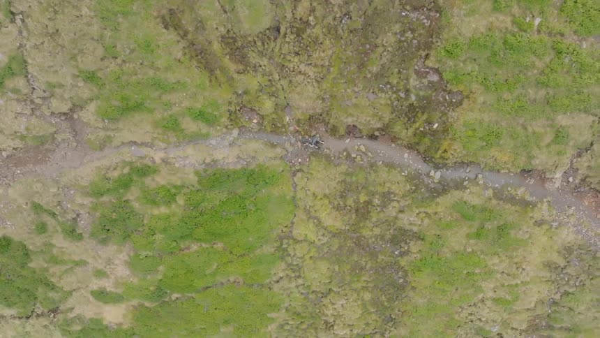 An aerial top-down view shows a mountain biker navigating a rugged, narrow trail through a vast green landscape