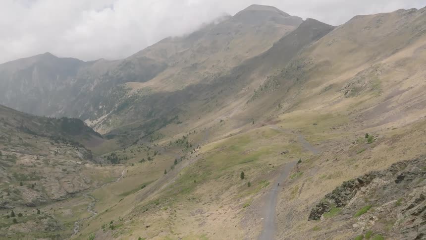 An epic aerial view follows a lone cyclist navigating a winding dirt road through a vast, remote mountain range