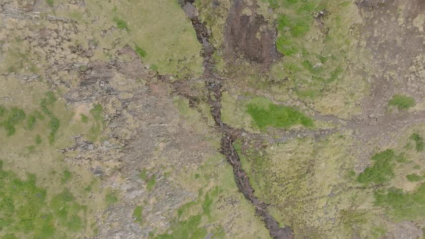 An aerial top-down shot slowly pulls back to reveal a small stream winding through the vast, green and rocky landscape of Iceland