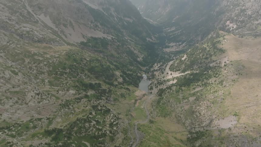 An aerial view slowly tilts up to reveal a vast, rugged mountain valley with a river and lake on a cloudy, hazy day